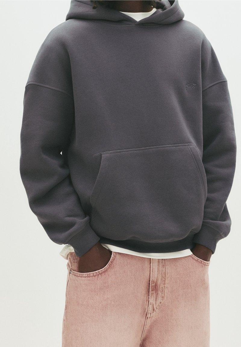 Personne portant un sweat à capuche gris foncé avec une poche avant et un jean rose pâle, les mains dans les poches du jean.