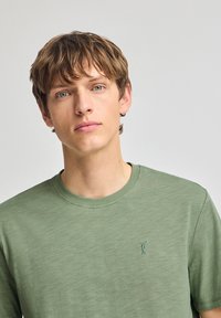 Grön kortärmad T-shirt gjord av mjukt tyg, med rund halsringning och en liten broderad design på bröstet i en liknande nyans.