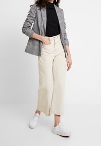 Blazer à carreaux gris avec une coupe ajustée, associé à un pantalon large crème et des baskets blanches. Sur un fond blanc uni.