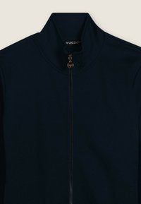 Chaqueta azul marino con cremallera y cuello alto, hecha de tela lisa. Cuenta con una cremallera de bronce y una etiqueta con el logo en el cuello.