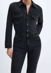 Mono de mezclilla negro con cuello de camisa, cierre de botones en la parte delantera, bolsillos en el pecho y mangas largas. Silueta ajustada, textura ligeramente desgastada.