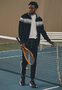 Svart velour treningsdress med grå striper, hvit t-skjorte, svarte joggesko, holder en oransje tennisracket, står nær et tennisnett.