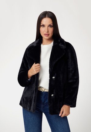 Giacca di pelliccia sintetica nera con un ampio colletto, due bottoni argentati e un taglio rilassato. Indossata sopra un maglione bianco a coste e jeans blu.