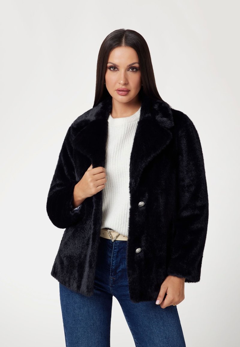 Giacca di pelliccia sintetica nera con un ampio colletto, due bottoni argentati e un taglio rilassato. Indossata sopra un maglione bianco a coste e jeans blu.