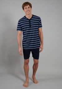 Marine gestreiftes Kurzarm-Henley-Shirt mit blauen und weißen Streifen, kombiniert mit marineblauen Shorts. Das Model steht barfuß vor einem grauen Hintergrund.