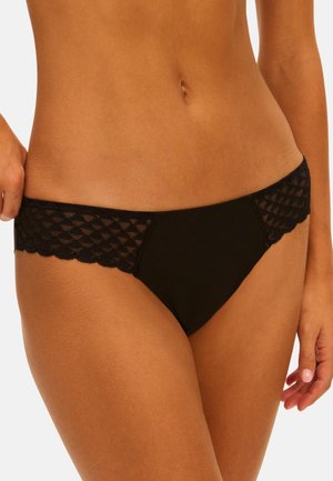 Frau richtet schwarze, spitzenbesetzte Bikini-Unterwäsche, zeigt Bauch und Hüften vor neutralem Hintergrund.