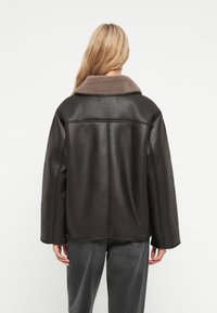 Brun kunstlæderjakke med en beige fleecekrave, oversized pasform og kontrasterende syninger. Bæres med grå løse jeans.