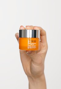 Oransje kremkrukke med sølvhette, merket "Clinique Superdefense," holdt i en hånd mot en hvit bakgrunn.