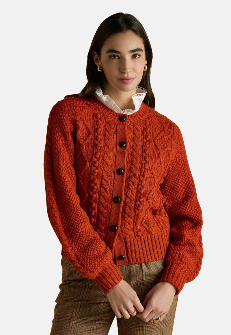 Oranžs adīts kardigāns ar apaļu kaklu, pogu aizdari un ribotu apakšmalu. Iespējas ar teksturētiem rakstiem un garām piedurknēm.