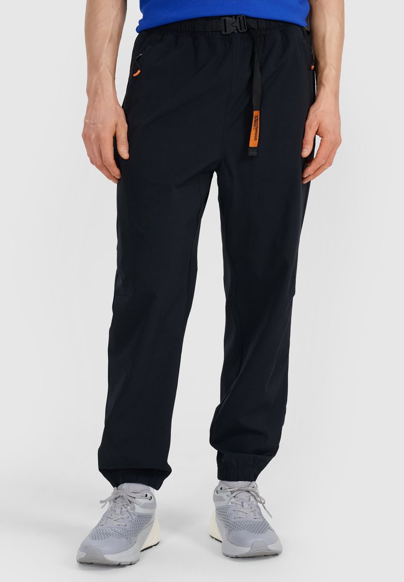 Personne portant un pantalon noir avec des poignets élastiques, une chemise bleue et des chaussures de sport grises, debout devant un fond uni.