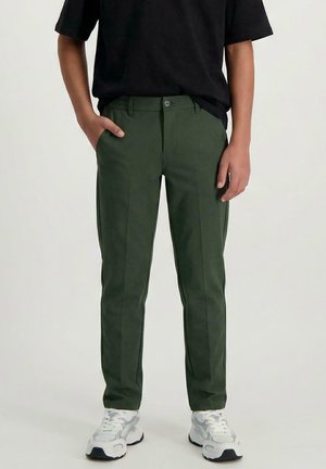 Persoon draagt donkergroene, op maat gemaakte broek met plooien, zwart shirt met korte mouwen, witte sneakers, staat met één hand in de zak.