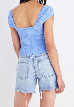 Vaaleansininen teksturoitu pusero, jossa on lyhyet pussihihat ja matala selkäosa, yhdistettynä resuiseen farkkushortseihin. Olkapäillä on avainreikäkuvio.