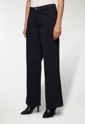 Donna che indossa jeans neri a vita alta e larghi, con stivali neri a punta e tacco, in piedi davanti a uno sfondo chiaro e uniforme.