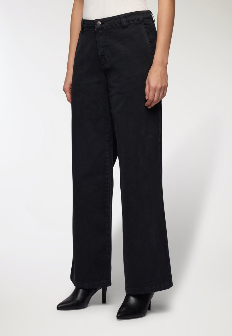 Donna che indossa jeans neri a vita alta e larghi, con stivali neri a punta e tacco, in piedi davanti a uno sfondo chiaro e uniforme.
