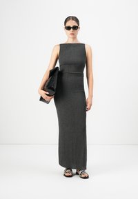 Kvinne som har på seg en ermeløs lang grå kjole, svarte solbriller, holder en stor svart clutch, og har på seg sandaler med slangeskinnsmønster, stående mot hvit bakgrunn.