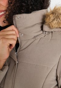 Chaqueta acolchada color beige con capucha forrada de suave pelo, cierre de cremallera, botones a presión y textura acolchada. Primer plano del cuello y mano agarrando la cremallera.