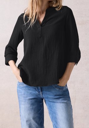 Femme portant un chemisier noir texturé à manches longues avec des poignets retroussés et un jean bleu clair délavé, les mains dans les poches.