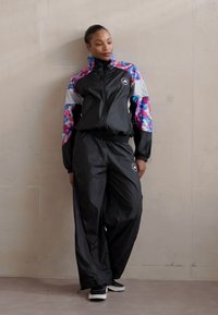 Survêtement noir avec une veste zippée présentant un imprimé floral coloré sur les épaules et sur les côtés, associé à un pantalon noir ample.