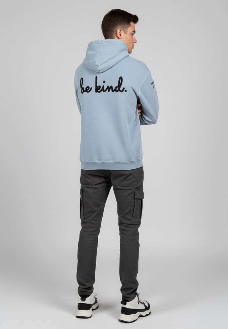 Ljust blå hoodie med en texturerad yta och "var snäll." i svart på baksidan. Kombinerad med mörkgrå cargobyxor och vita sneakers.