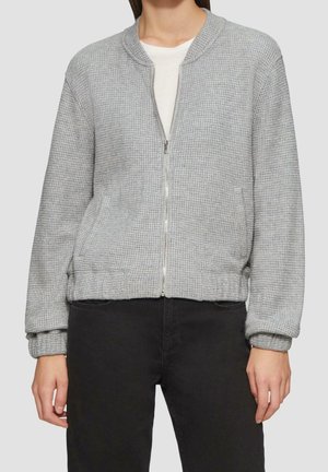 Kvinde iført en lysegrå waffle-strikket jakke med lynlås over en hvid skjorte og sorte bukser, stående mod en neutral baggrund.