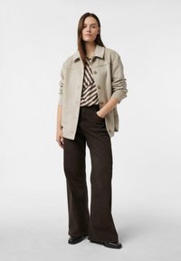 Eine Frau steht mit einer Hand in der Tasche, trägt eine beige, knöpfbare Jacke, ein gestreiftes Oberteil in Beige und Braun, weite dunkle Hose und schwarze Schuhe.