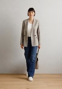 Blazer a righe grigie e bianche con tasche frontali, abbinato a una camicia bianca e jeans blu. Il modello tiene una borsa grigia. Sneakers bianche completano l'outfit.