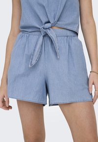 Vaalean siniset farkkushortsit, joissa on joustava vyötärö ja solmittava yksityiskohta edessä. Ominaisuuksiin kuuluu rento istuvuus ja sileä rakenne.