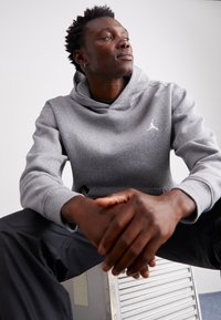 Szürke kapucnis pulóver elülső zsebből és nagy kapucnis kialakításból, fehér Jumpman logóval. Fekete nadrággal viselve, kezei a térdén pihennek.