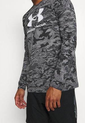 Mand iført en grå og sort digital camouflage Under Armour langærmet skjorte og sorte shorts, stående mod en hvid baggrund.
