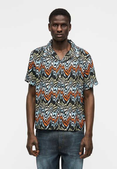 Άνδρας που φοράει κοντομάνικο πουκάμισο με πολύχρωμο ζιγκ-ζαγκ σχέδιο και σκούρα μπλε τζιν, στέκεται μπροστά σε λευκό φόντο.