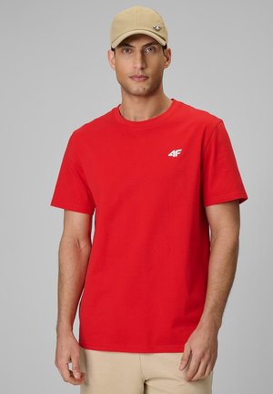 Mężczyzna w czerwonym t-shircie z krótkim rękawem i małym białym logo na piersi, beżowe spodnie i beżowa czapka z daszkiem, stojący na tle szarego tła.