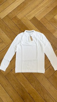 Camisa branca de mangas longas com colarinho clássico, textura suave e etiquetas anexadas, deitada em plano sobre um chão de madeira com padrão em espinha de peixe.