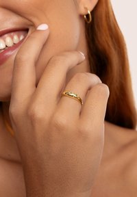 Anello d'oro con una finitura liscia e lucida, caratterizzato da un design sottile e da una piccola pietra di accentazione rosa. La mano poggia vicino al viso.