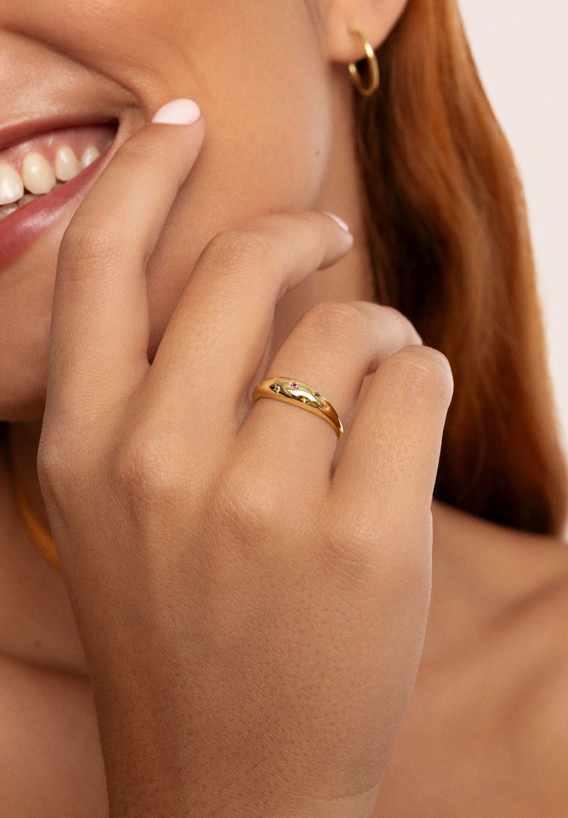 Anello d'oro con una finitura liscia e lucida, caratterizzato da un design sottile e da una piccola pietra di accentazione rosa. La mano poggia vicino al viso.