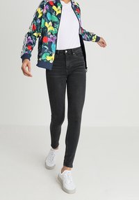 Persona caminando vistiendo jeans ajustados negros, zapatillas blancas, camiseta blanca y una chaqueta de patrón floral colorido.