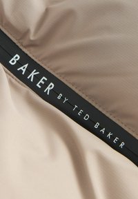Beige Stoff mit glatter Textur, auf dem ein schwarzes Etikett angebracht ist. Das Etikett trägt den Text "BAKER BY TED BAKER" in weißer, mittig angeordneter Schrift.