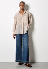Camisa de botón a rayas beige claro con un bolsillo en el pecho, combinada con unos pantalones vaqueros anchos de color azul. Los vaqueros tienen un acabado desgastado y una costura simple.