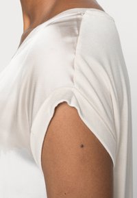 T-shirt beige chiaro con scollo a V e maniche corte arrotolate, realizzato in un tessuto morbido e liscio che scende leggermente.