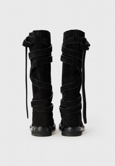 Para czarnych zamszowych kozaków za kolano z owijającymi paskami i klamrami, pokazanych z tyłu na jasnym białym tle.