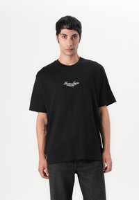 T-shirt en coton noir à manches courtes, col rond et logo "Guess Jeans" brodé en blanc sur la poitrine. Unicolore, coupe décontractée.