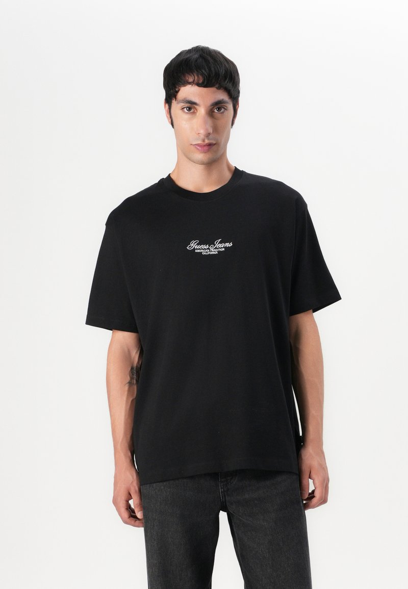 T-shirt en coton noir à manches courtes, col rond et logo "Guess Jeans" brodé en blanc sur la poitrine. Unicolore, coupe décontractée.