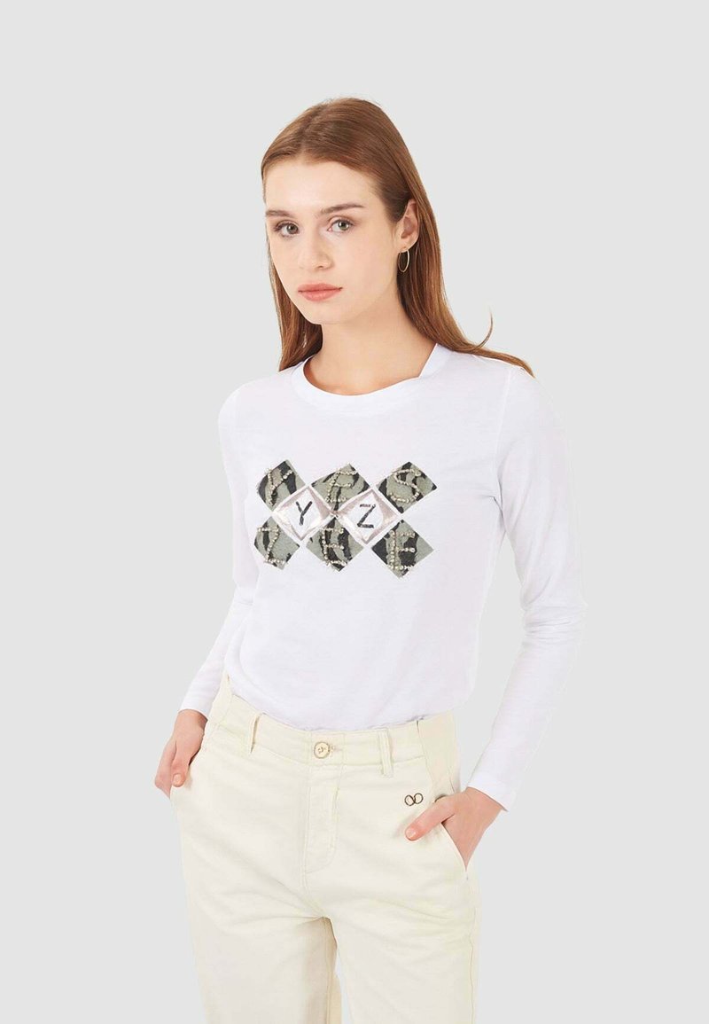 Camicia bianca a maniche lunghe con una grafica a forma di diamante con un motivo mimetico e lettere nere. Abbinata a pantaloni beige chiaro.