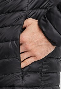 Giacca a vento nera con texture trapuntata, dotata di una tasca laterale con zip e una mano appoggiata all'interno, che mostra una finitura in tessuto liscia.