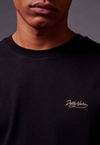 T-shirt en coton noir avec col rond et logo brodé beige sur la poitrine gauche. Texture lisse avec une coupe classique.