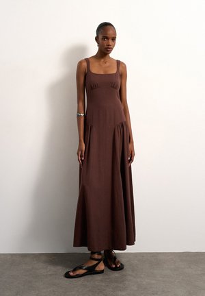 Vrouw in een mouwloze bruine maxi-jurk met een vierkante halslijn en zwarte sandalen met enkelriempjes, staand tegen een witte muur.