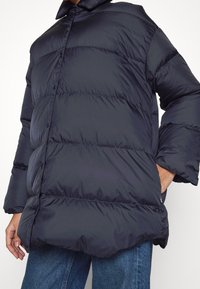 Navy Blousonjacke mit hohem Kragen, gestepptem Design und Druckknopfverschlüssen. Hergestellt aus glattem, glänzendem Material und im entspannten Schnitt.