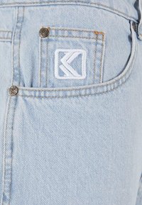 Svetlo modra denim tkanina vsebuje zadnji žep z belim vezenim logotipom, bakrene zakovice in vidne šivane detajle.