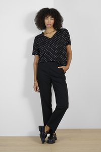 Schwarze Bluse mit weißen Punkten und V-Ausschnitt, kombiniert mit schwarzen, taillierten Hosen und klobigen schwarzen Schuhen, an einem Model vor einem weißen Hintergrund.