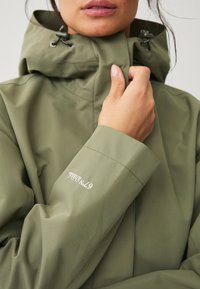 Chaqueta impermeable verde oliva con capucha con cordón, que presenta una textura suave y un detalle del logo en la manga. Primer plano de una mano agarrando el cuello.
