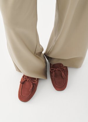 Person iført beige bukser med vide ben og rustrøde mokkasiner i ruskind med kvaster på et hvidt gulv.